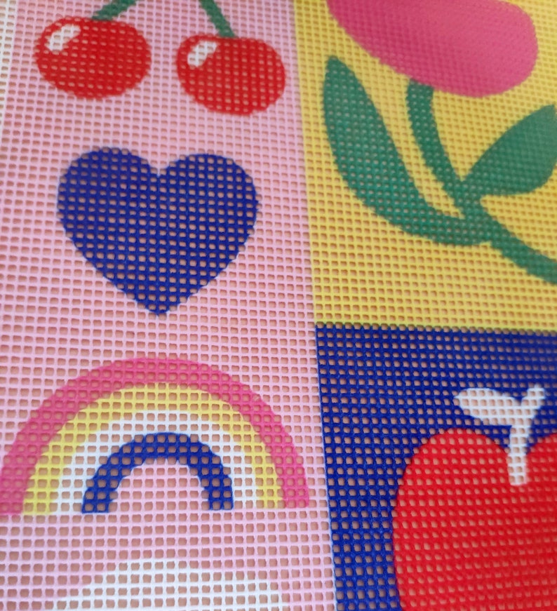 Peut inclure: Une toile de point de croix color&eacute;e avec un motif r&eacute;p&eacute;t&eacute; de cerises, un c&oelig;ur bleu, un arc-en-ciel, une fleur et une pomme rouge. Le fond est divis&eacute; en carr&eacute;s roses et jaunes, avec une texture quadrill&eacute;e.