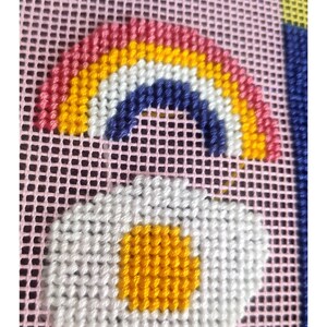 Peut inclure: Motif de point de croix repr&eacute;sentant un arc-en-ciel avec des rayures rouges, orange, jaunes et blanches, et une marguerite blanche avec un centre jaune. Le motif est sur un fond en maille rose avec des formes g&eacute;om&eacute;triques bleues et jaunes.