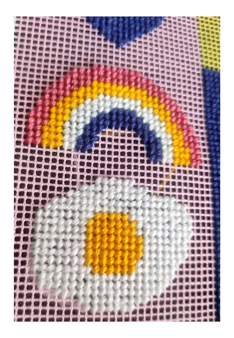 Peut inclure: Motif de point de croix repr&eacute;sentant un arc-en-ciel avec des rayures rouges, orange, jaunes et blanches, et une marguerite blanche avec un centre jaune. Le motif est sur un fond en maille rose avec des formes g&eacute;om&eacute;triques bleues et jaunes.