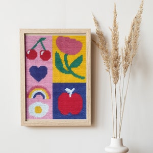 Peut inclure: Une &oelig;uvre d'art au point de croix encadr&eacute;e, aux couleurs vives, repr&eacute;sentant une cerise, un c&oelig;ur, un arc-en-ciel, une fleur, une pomme et un &oelig;uf. L'&oelig;uvre est encadr&eacute;e dans un cadre en bois clair.