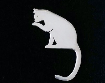 May include: A silver-colored metal cat figurine in a sitting position, with its tail curled around its body. The cat is depicted grooming itself with its paw.