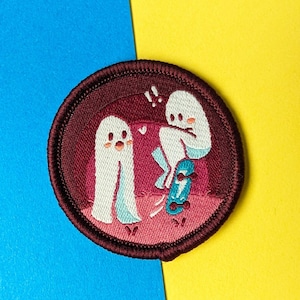 Może przedstawiać: Naszywka haftowana przedstawiająca dwa białe duchy, jeden stojący, a drugi jeżdżący na deskorolce, na bordowym tle z różowymi akcentami. Naszywka ma czarną ramkę.