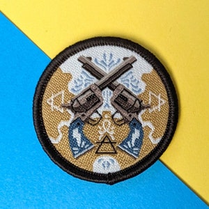 以下が含まれることがあります： 白い背景と金色の縁取りのある丸い刺繍パッチ。パッチの中央には、交差した2丁の拳銃があり、その周りを青と白のデザインが囲んでいます。パッチは黒い縁取りをしています。