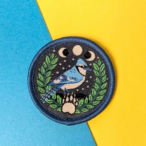 May include: An embroidered patch featuring a blue jay bird against a dark blue background, with green leaves, a white paw print and three white moons. The patch has a starry night sky design.