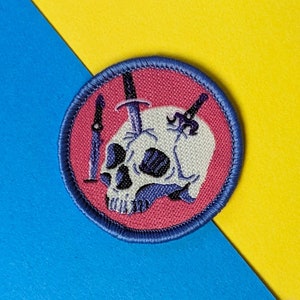 以下が含まれることがあります： 白の頭蓋骨とそれを貫く3本の黒い短剣が描かれた、ピンクとブルーの刺繍パッチです。