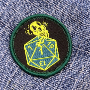 May include: A green and black embroidered patch with a yellow die and a yellow ghost-like creature. The die has the number 1 on it and the text "D1" on the bottom.