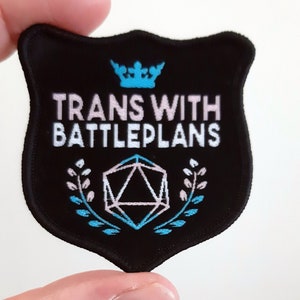 Può includere: Una toppa ricamata nera con una corona blu, testo bianco che recita "TRANS WITH BATTLEPLANS", una forma geometrica rosa e blu e disegni floreali blu e rosa.