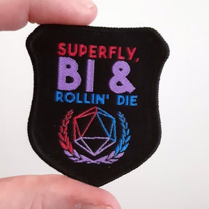 May include: A black embroidered patch with the text "SUPERFLY, BI & ROLLIN' DIE" in purple and red lettering. A purple, blue, and red dice is in the centre of the patch with a wreath of purple leaves around it.