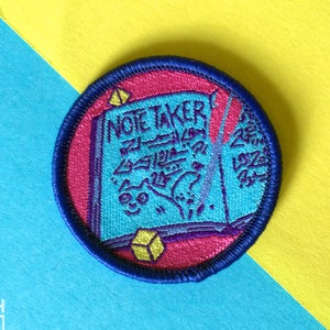 Peut inclure: Un patch brodé bleu et rose avec un livre fermé et le texte "NOTE TAKER" en violet. Le livre est rempli de gribouillis violets et un cube jaune se trouve devant le livre.