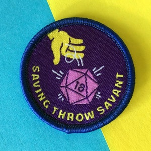 以下が含まれることがあります： 青い縁取りのある丸い紫色のパッチ。18の数字が入ったピンク色の20面ダイスの上に黄色の手があります。「SAVING THROW SAVANT」の文字がダイスの周りに黄色で書かれています。青と黄色の背景。