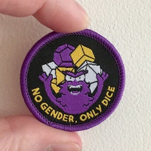 May include: Purple and yellow embroidered patch with a cartoon monster holding dice. The text on the patch reads "No Gender, Only Dice".