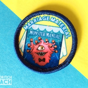 Op de afbeelding: Een blauwe en gele geborduurde patch met de tekst "Marriage Material" en een monster met meerdere ogen en een strikje. Het monster houdt een boek vast met de tekst "Monster Manual".