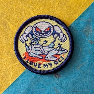 May include: A circular embroidered patch with a dark blue border and a pale yellow background. It features a white creature with red eyes and sharp teeth standing over a fallen figure, with the text I LOVE MY OCS printed in black along the bottom.