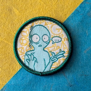 May include: A circular embroidered patch featuring a light blue alien character with large eyes and a speech bubble. The background of the patch is white with a yellow geometric pattern. The patch has a dark green stitched border edge.