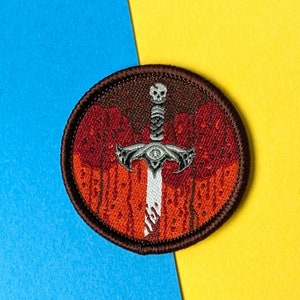 May include: A brown embroidered patch with a white sword with a skull at the top and an eye in the middle of the blade. The sword is surrounded by red and orange flames.