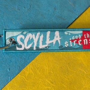 May include: A rectangular keychain tag with the text "SCYLLA and the sirens" in white lettering. The tag has a teal background with a wave design and a red star. A silver key ring is attached.