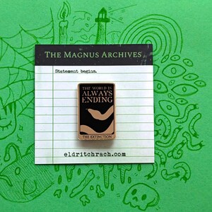 Pu&ograve; includere: Spilla rettangolare con la scritta "THE WORLD IS ALWAYS ENDING" su sfondo nero con un disegno beige. La spilla &egrave; su una carta bianca con la scritta "THE MAGNUS ARCHIVES" e "Statement begins." Lo sfondo &egrave; verde con scarabocchi verdi.