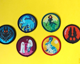 May include: Six embroidered patches featuring various creatures and designs. The patches are circular and have a black outline. The designs include a blue plesiosaur, a green humanoid figure, a black bat, two white ghosts, a blue rabbit, and a black rabbit with the text "HEWWO".