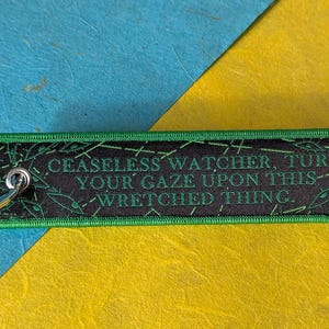 Puede incluir: Un llavero rectangular bordado en negro y verde con el texto "CEASELESS WATCHER, TURN YOUR GAZE UPON THIS - WRETCHED THING." El llavero tiene un llavero plateado adjunto. El fondo es azul y amarillo.