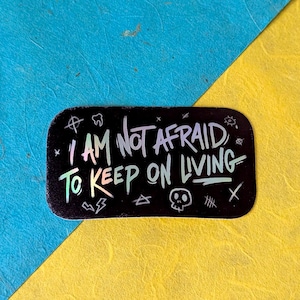 Puede incluir: Una pegatina rectangular negra con esquinas redondeadas, con el texto "I AM NOT AFRAID TO KEEP ON LIVING" en letras blancas e iridiscentes. La pegatina tiene varias ilustraciones pequeñas y está sobre un fondo azul y amarillo.