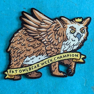 Puede incluir: Parche bordado con un híbrido de búho y oso de dibujos animados con alas, una corona y el texto "FAT OWLBEAR WEEK CHAMPION" en una pancarta. El parche es marrón, beige y blanco con contornos negros. El fondo es azul.