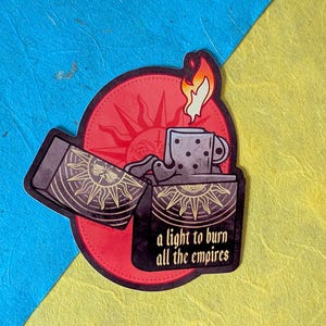 May include: A sticker depicting a stylized lighter with a flame, a cup, and the text "a light to burn all the empires." The design includes a red sunburst and intricate patterns in gold and black.