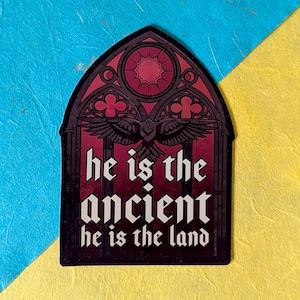 Peut inclure: Autocollant bordeaux et noir avec un motif de fenêtre gothique, un oiseau stylisé et le texte blanc "he is the ancient he is the land" en lettres gothiques. L'autocollant est sur un fond bleu et jaune.