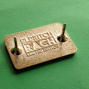 Pu&ograve; includere: Spilla rettangolare color oro con la scritta in rilievo "ELDRITCH RACH LIMITED EDITION". Sono visibili due fermi per spilla. La spilla &egrave; posta su uno sfondo verde.