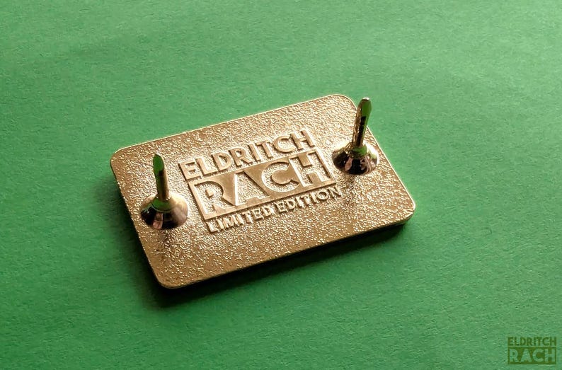 Pu&ograve; includere: Spilla rettangolare color oro con la scritta in rilievo "ELDRITCH RACH LIMITED EDITION". Sono visibili due fermi per spilla. La spilla &egrave; posta su uno sfondo verde.