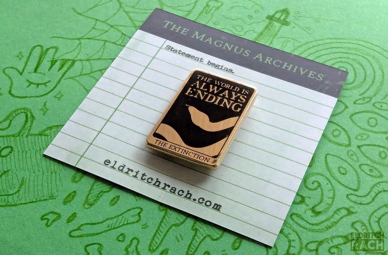 Pu&ograve; includere: Spilla rettangolare con la scritta "THE WORLD IS ALWAYS ENDING" su sfondo nero, con bordo dorato. La spilla &egrave; su un blocco note con la scritta "THE MAGNUS ARCHIVES" e "Statement begins." Lo sfondo &egrave; verde con scarabocchi.