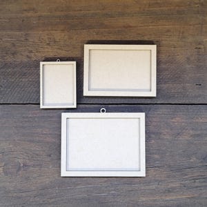 May include: Three light beige rectangular frames made of unfinished wood material. One frame is oriented vertically, while two frames are oriented horizontally. Each frame features a small circular loop at the top for hanging purposes.