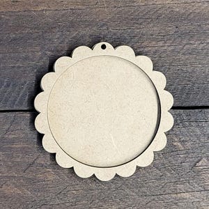 May include: A round, unfinished wooden craft blank with a scalloped edge and a hole at the top for hanging. The center is open, creating a frame-like appearance. The wood has a light brown color.