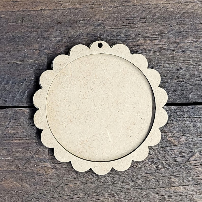 May include: A round, unfinished wooden craft blank with a scalloped edge and a hole at the top for hanging. The center is open, creating a frame-like appearance. The wood has a light brown color.