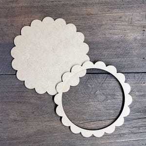 May include: Two unfinished wooden craft pieces with a scalloped edge. One is a solid circle, and the other is a ring with the same scalloped design. The pieces are light brown and sit on a dark wood grain surface.