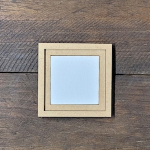 May include: A small, square, light-colored wooden frame with a white square canvas insert. The frame has a double border design. The item is set against a dark brown, wooden background.