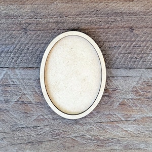 May include: An oval-shaped wooden craft blank with a thin black border. The unfinished wood has a light beige color and is set against a rustic wooden background. This craft supply is ideal for DIY projects and home decor.