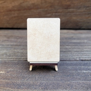 May include: A blank, light brown, rectangular sign with rounded corners is standing on a small wooden easel. The sign is made of wood and has a natural, unfinished look.