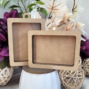 May include: Two rectangular wooden frames with a cutout center and a small circular hole at the top for hanging. The frames are light brown and sit on a white surface with decorative flowers in the background. The frames are of different sizes.
