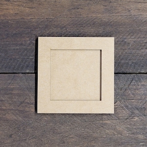 May include: A square, light brown picture frame with a recessed center, resting on a dark wooden surface. The frame is made of a light-colored material, possibly wood or cardboard, and has a simple, clean design. The frame is empty.