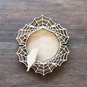 May include: A decorative wooden frame with a spiderweb design and a raven silhouette. The frame has a circular opening and is made of light-colored wood. The raven is perched on the frame's edge, adding a spooky touch. Perfect for Halloween decor.