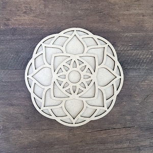 May include: A wooden mandala with a intricate design featuring multiple layers of overlapping circles and flower-like shapes. The mandala is cut out of a single piece of wood and has a natural, unpainted finish.