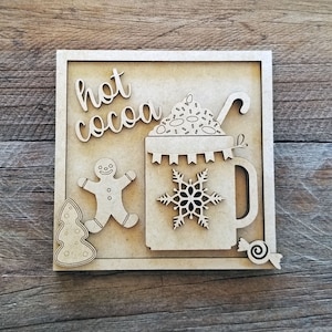 May include: A wooden sign with the words "hot cocoa" in a decorative font. The sign features a cutout of a mug of hot cocoa with whipped cream and sprinkles, a gingerbread man, a candy cane, a snowflake, a Christmas tree, and a candy swirl.