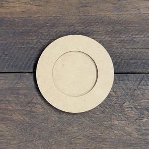May include: A round, unfinished wooden coaster with a recessed center. The coaster is light brown and sits on a dark brown, textured wooden surface. The coaster's design is simple and minimalist.