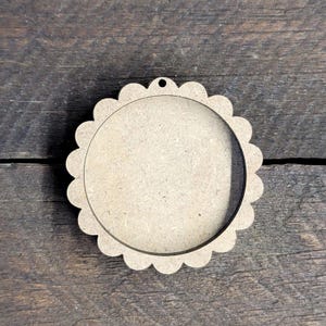 May include: A round, unfinished wooden craft blank with a scalloped edge and a small hole at the top for hanging. The center is open, creating a frame-like appearance. The wood has a natural, light brown color.