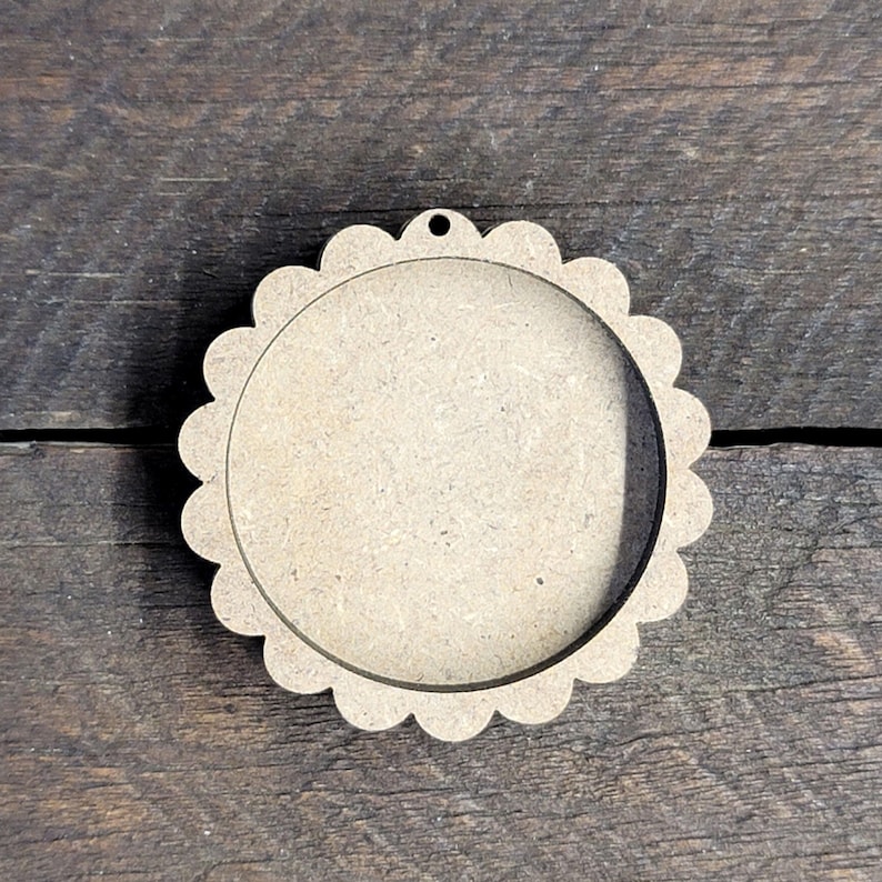 May include: A round, unfinished wooden craft blank with a scalloped edge and a small hole at the top for hanging. The center is open, creating a frame-like appearance. The wood has a natural, light brown color.