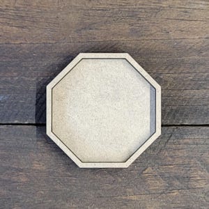 May include: An octagonal wooden coaster with a light brown surface and a dark brown border. The coaster is set against a dark wood grain background. This coaster is suitable for drinks.
