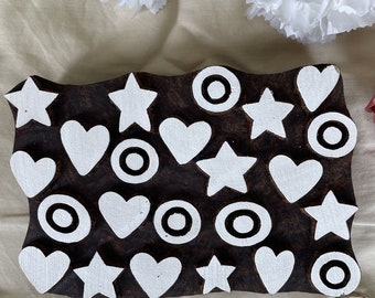 May include: A rectangular dark brown wooden printing block featuring a repeating pattern of white painted shapes including five-pointed stars, hearts, and concentric circles. The surface of the block is flat with raised carved motifs.