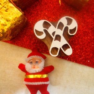 May include: A wooden printing block carved in the shape of two crossed candy canes with a natural brown finish, positioned alongside a small Santa Claus figurine wearing a red suit with a white beard and a gold belt, and gold gift boxes.