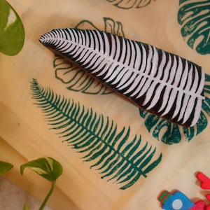 Puede incluir: Un sello de madera con un diseño de hoja de helecho, con tinta blanca y negra, descansa sobre una tela amarilla estampada con patrones de hojas verdes. Recortes decorativos de madera de criaturas marinas están esparcidos alrededor de la tela.