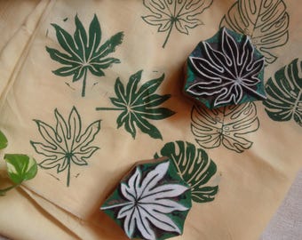 May include: Two wooden printing blocks with carved leaf designs resting on a light yellow fabric. The fabric features multiple dark green stamped leaf patterns, including palm-like and monstera leaf shapes, created by the blocks.